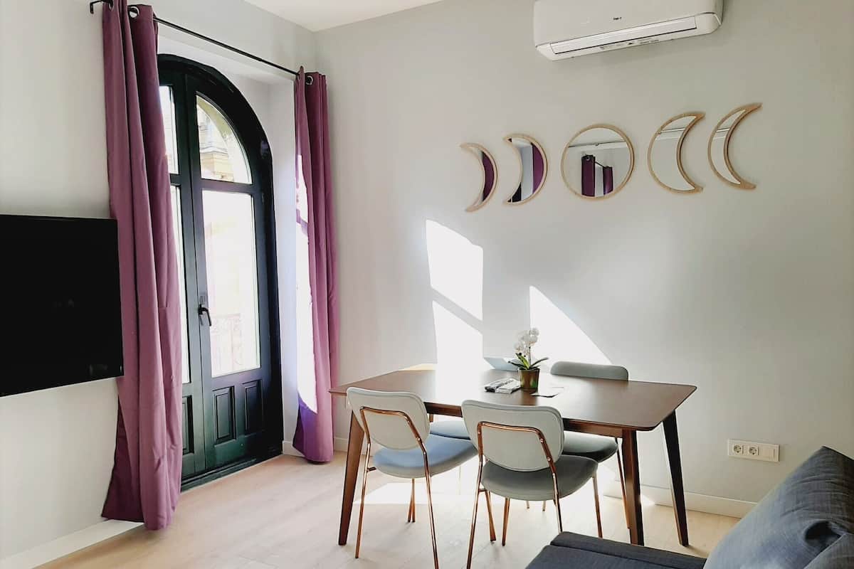 A modern living room with a dining area, featuring a large window with curtains, a flat-screen TV, a dining table with chairs, and decorative wall mirrors.