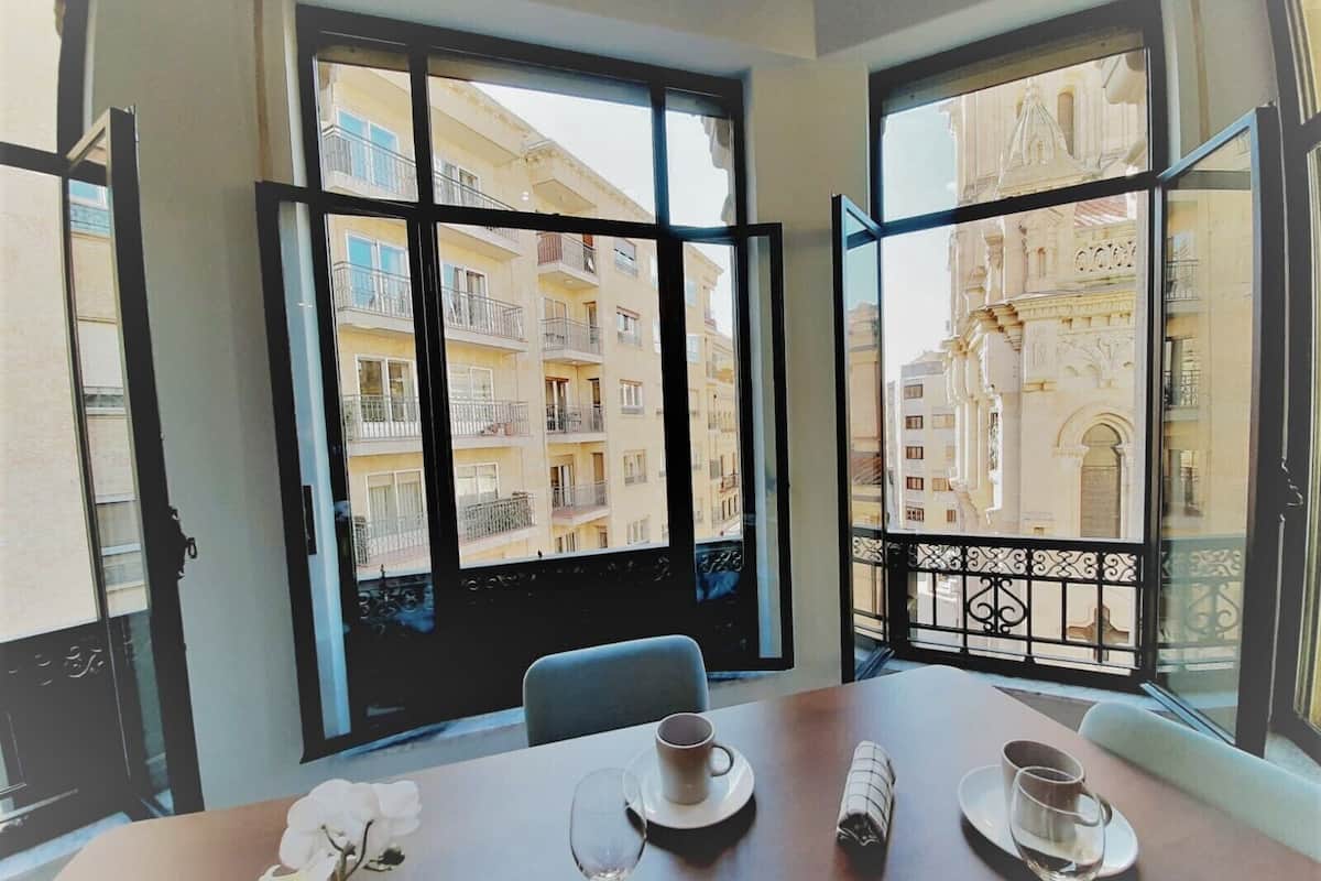 A dining area with a table set for two, a view of a building with balconies, and a balcony railing.