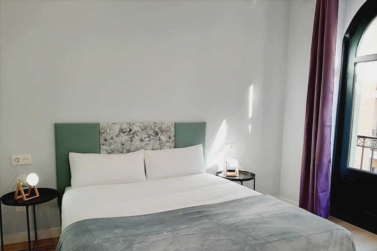 A hotel room with a bed, a nightstand, a lamp, and a view of the outside.