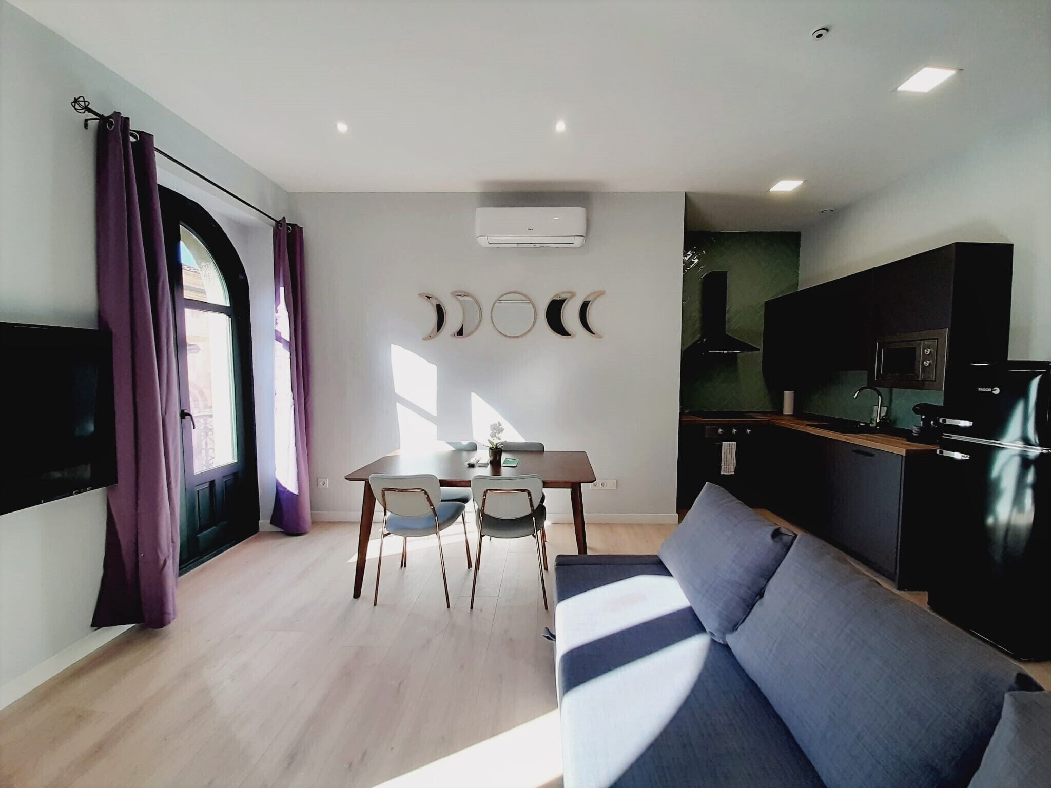A modern living room with a sofa, dining table, and chairs. There is a kitchen area with a stove, microwave, and refrigerator.
