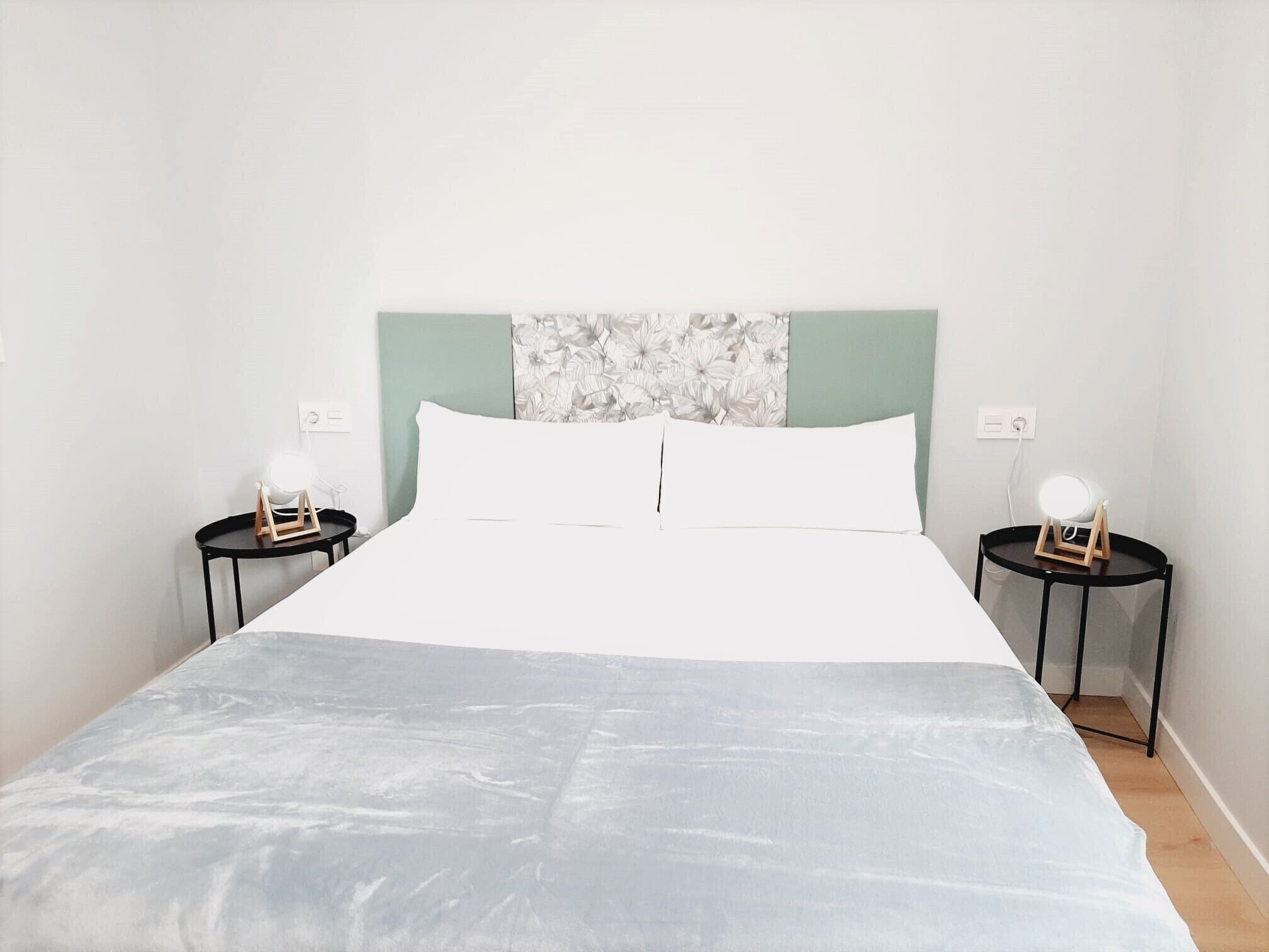 A neatly made bed with a white and grey bedspread, two bedside tables with lamps, and a wall with a floral pattern.