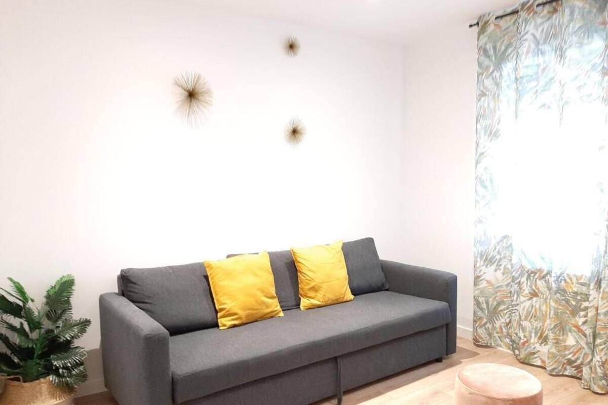 A grey sofa with two yellow cushions, a potted plant, and a round ottoman in a room with a light-colored floor and a curtain with a tropical leaf pattern.