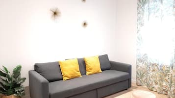 Un sofá gris con dos cojines amarillos, una planta en maceta y un otoman redondo en una habitación con piso de colores claros y una cortina con estampado de hojas tropicales.