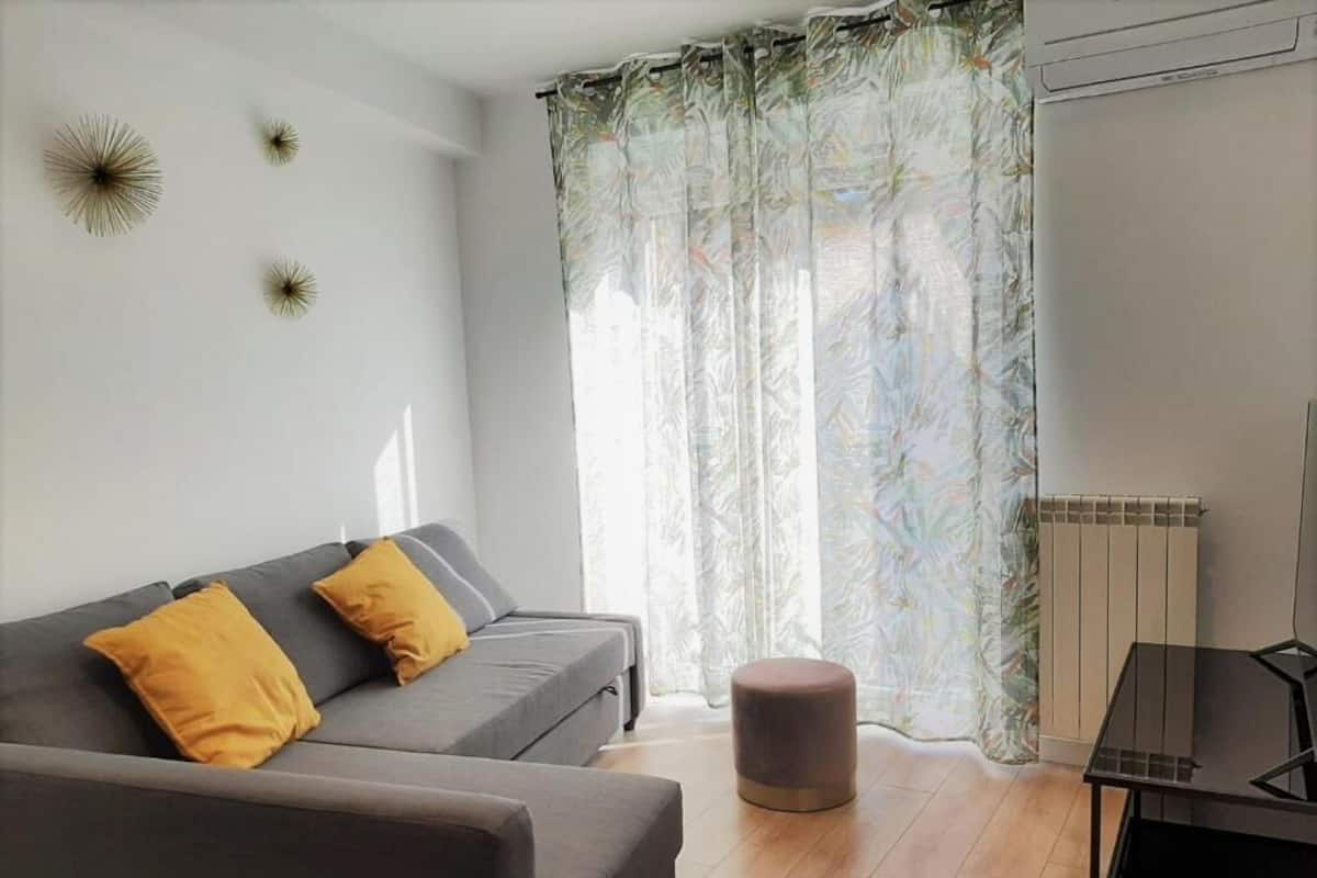 A modern living room with a grey sofa, two mustard pillows, a round ottoman, and a black coffee table.