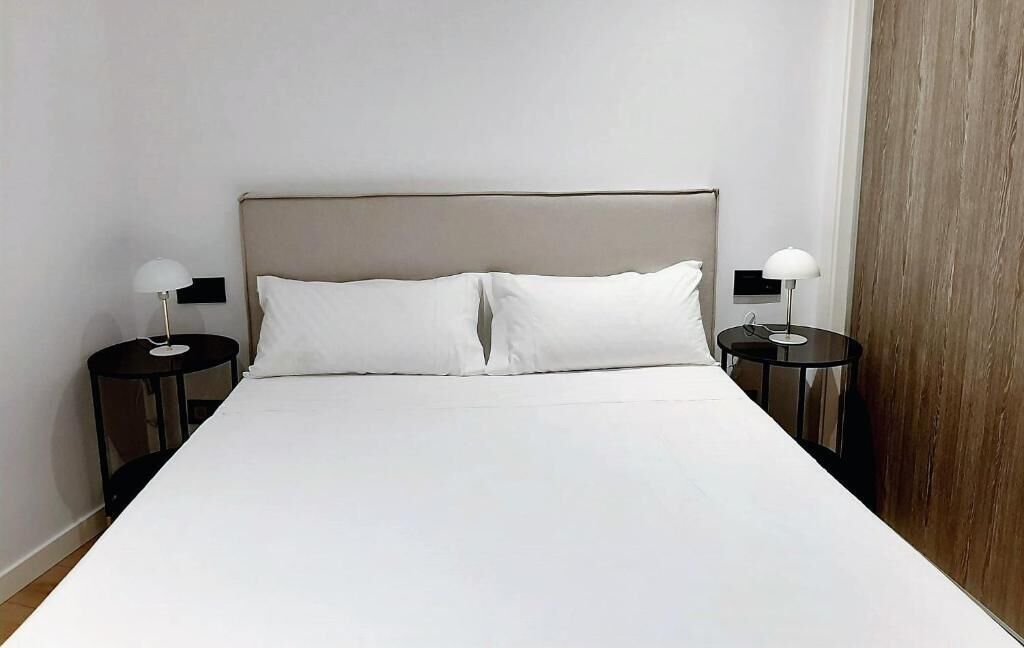 A neatly made bed with white linens and a wooden headboard. There are two bedside tables with lamps on either side of the bed.