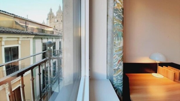 A balcony with a view of historic buildings, a wooden table with a lamp, and a patterned curtain. - Apartamentos Celestina by gaiarooms (Salamanca)