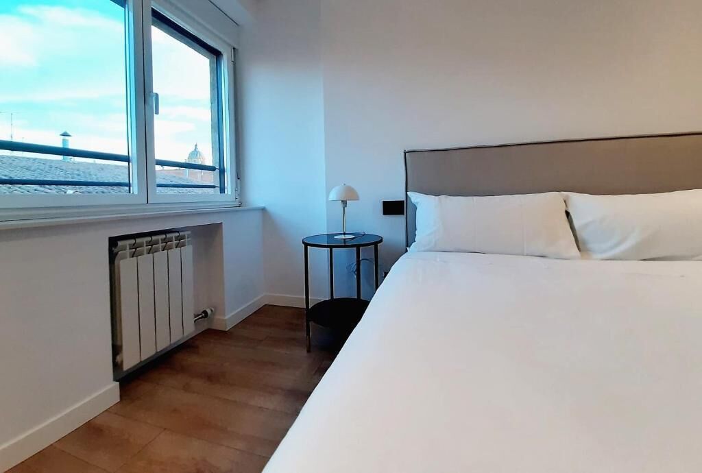 A hotel room with a large bed, a window with a view of a building, a small table with a lamp, and a radiator.