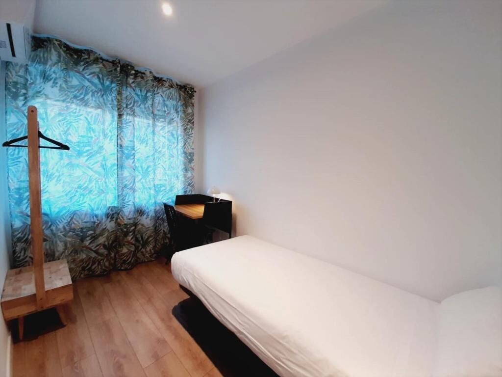 A small, tidy room with a single bed, a wooden hanger, a desk with a chair, and a window with patterned curtains.