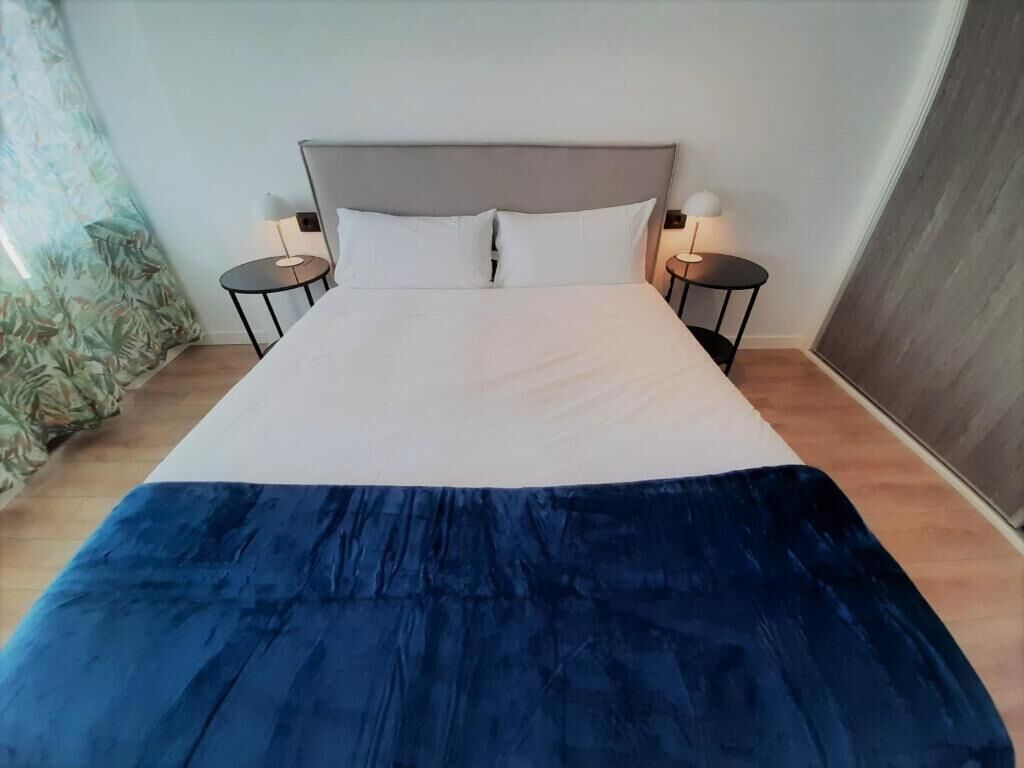 Una cama con cabecera gris, dos mesitas de noche con lámparas y una colcha azul.