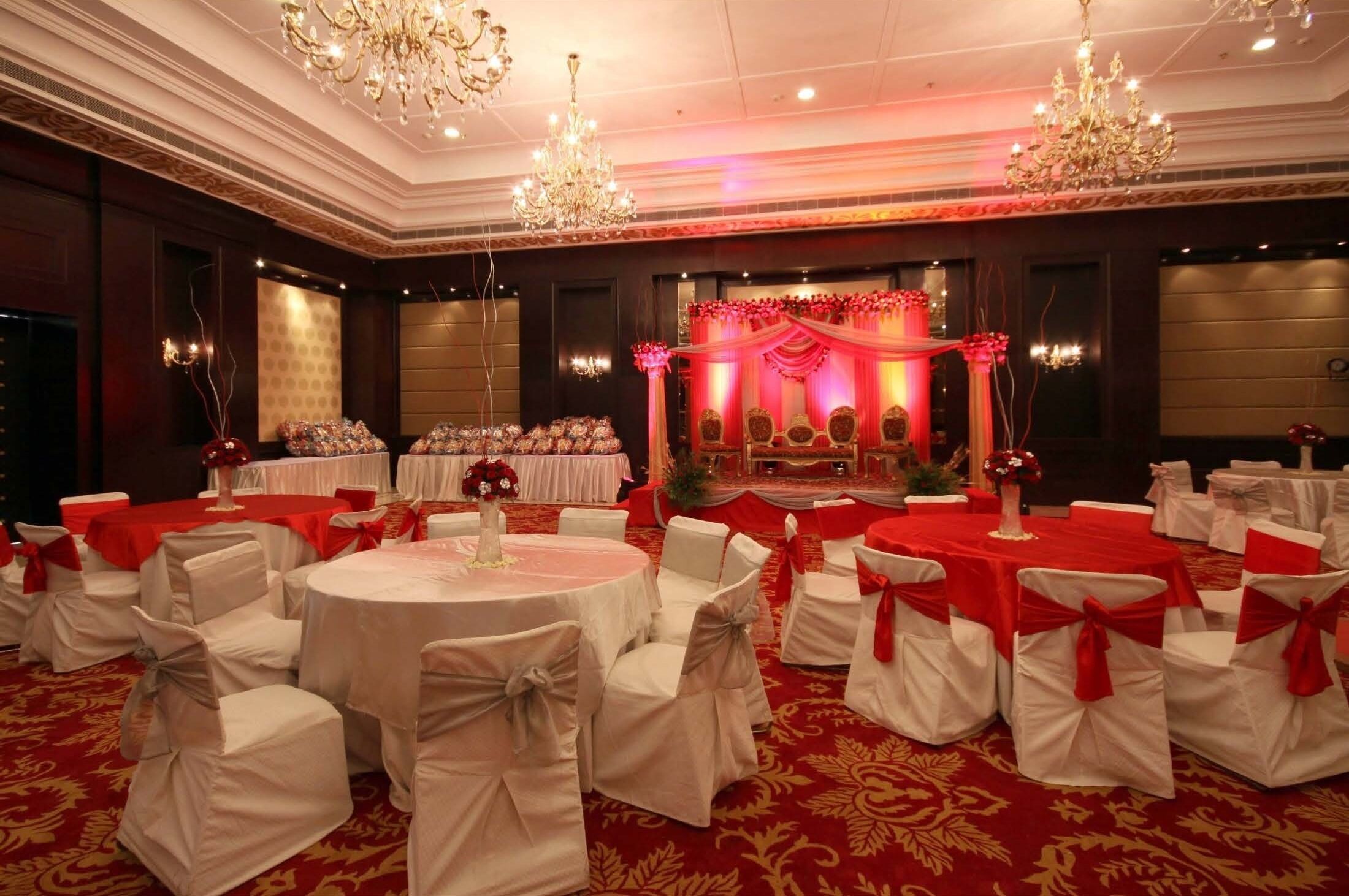 A banquet hall with round tables, white chairs with red sashes, chandeliers, and a stage with red drapery.
