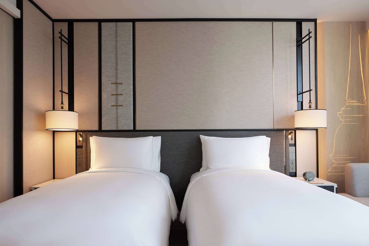 A hotel room with two beds, a nightstand with a lamp, and a wall-mounted control panel.