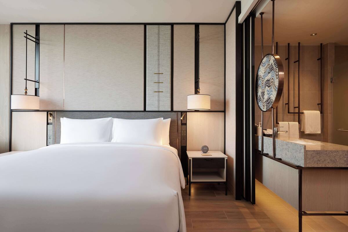 A modern hotel room with a large bed, bedside lamps, a small table with a clock, a mirror, and a bathroom with a sink and towel rack.