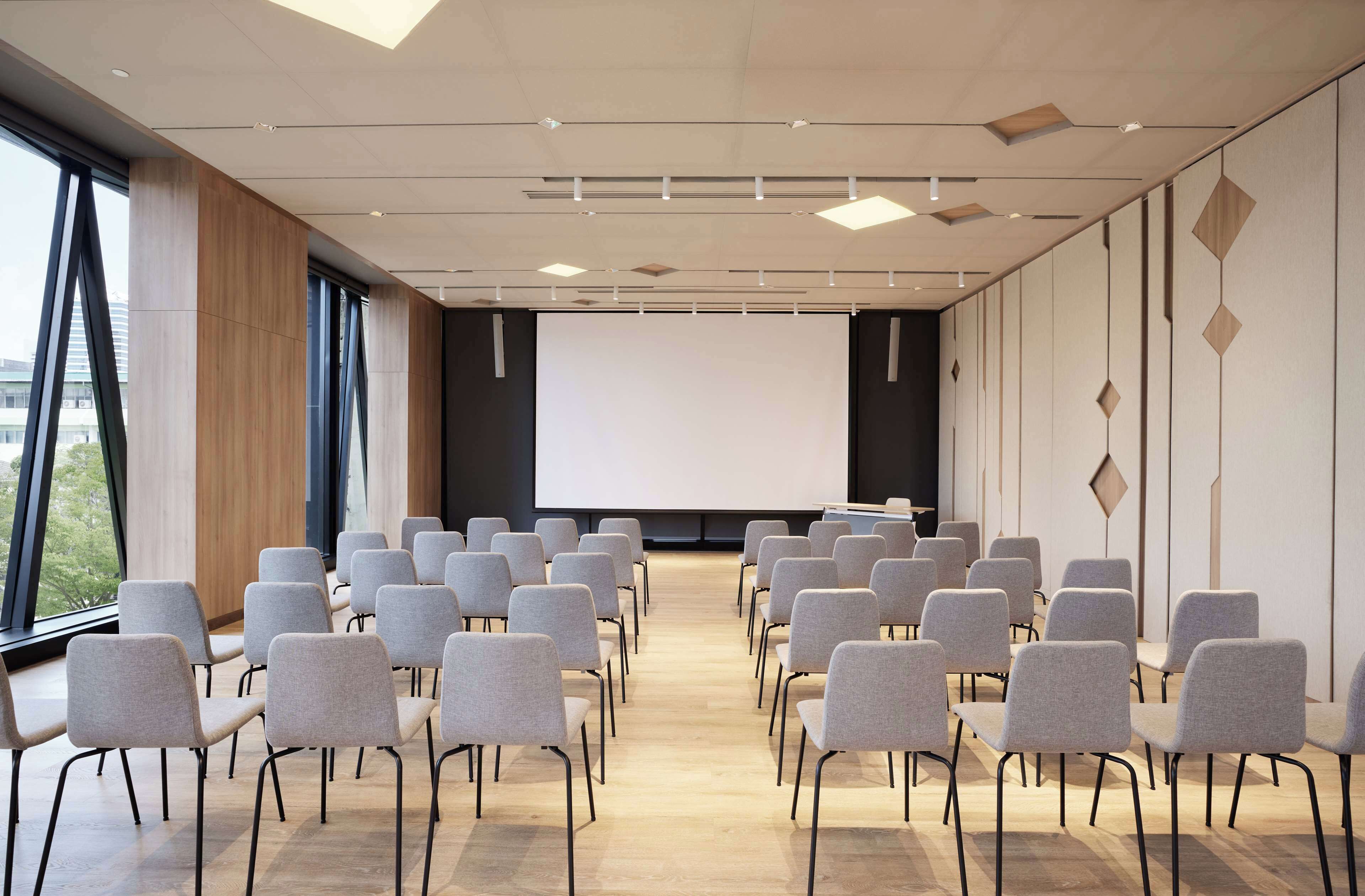 A conference room with rows of chairs facing a projection screen, large windows, and a modern ceiling design.
