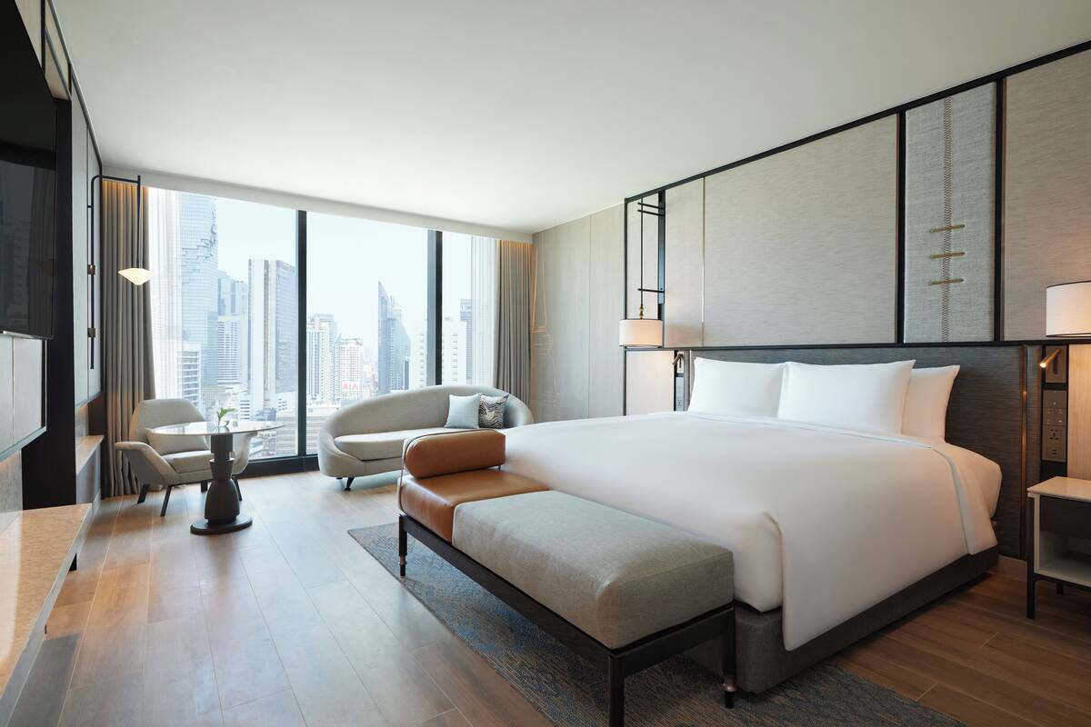 A modern hotel room with a large bed, a seating area with a table, and a view of the city skyline.