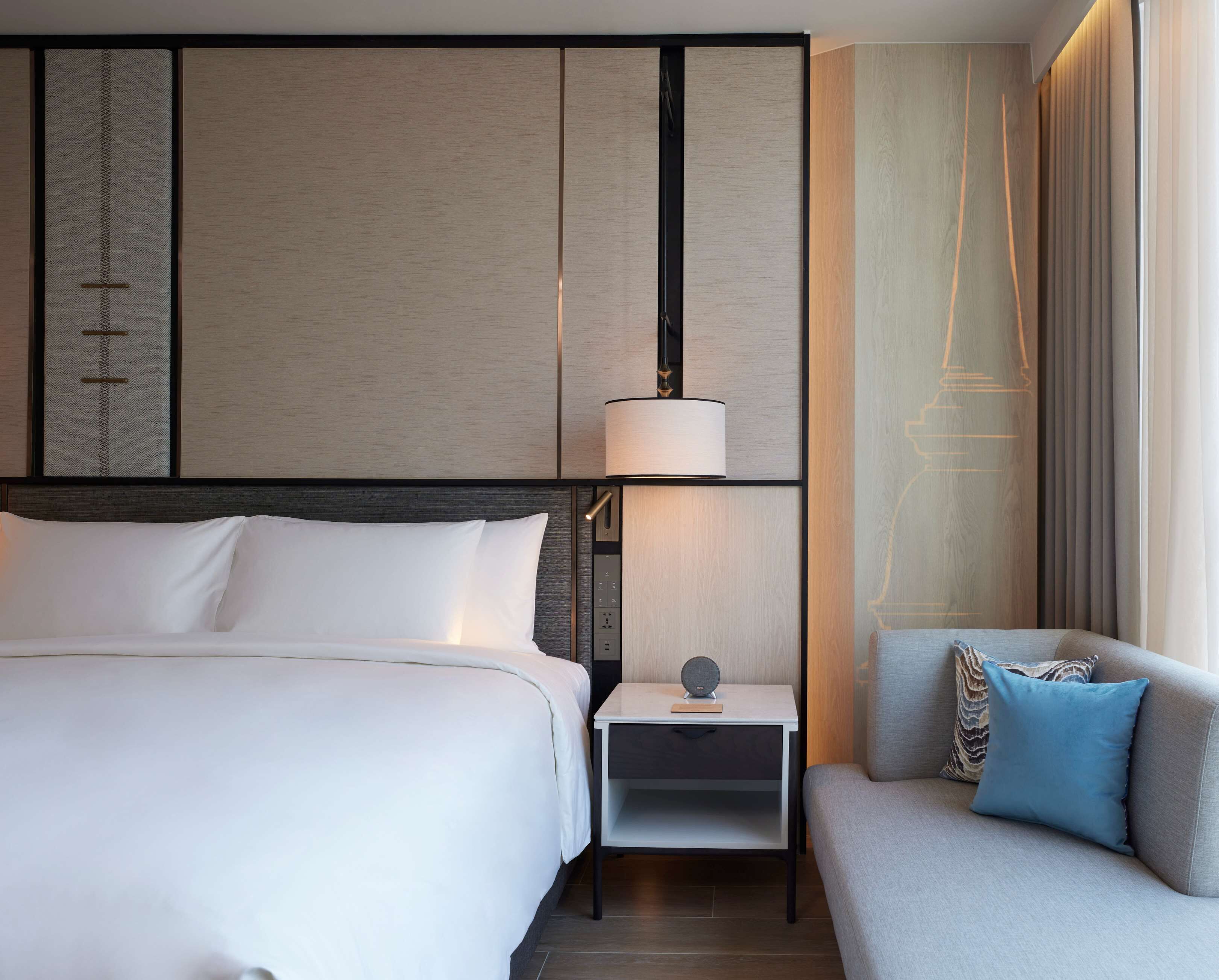 A modern hotel room with a large bed, a bedside table with a lamp, a small table with a speaker, and a sofa with pillows.