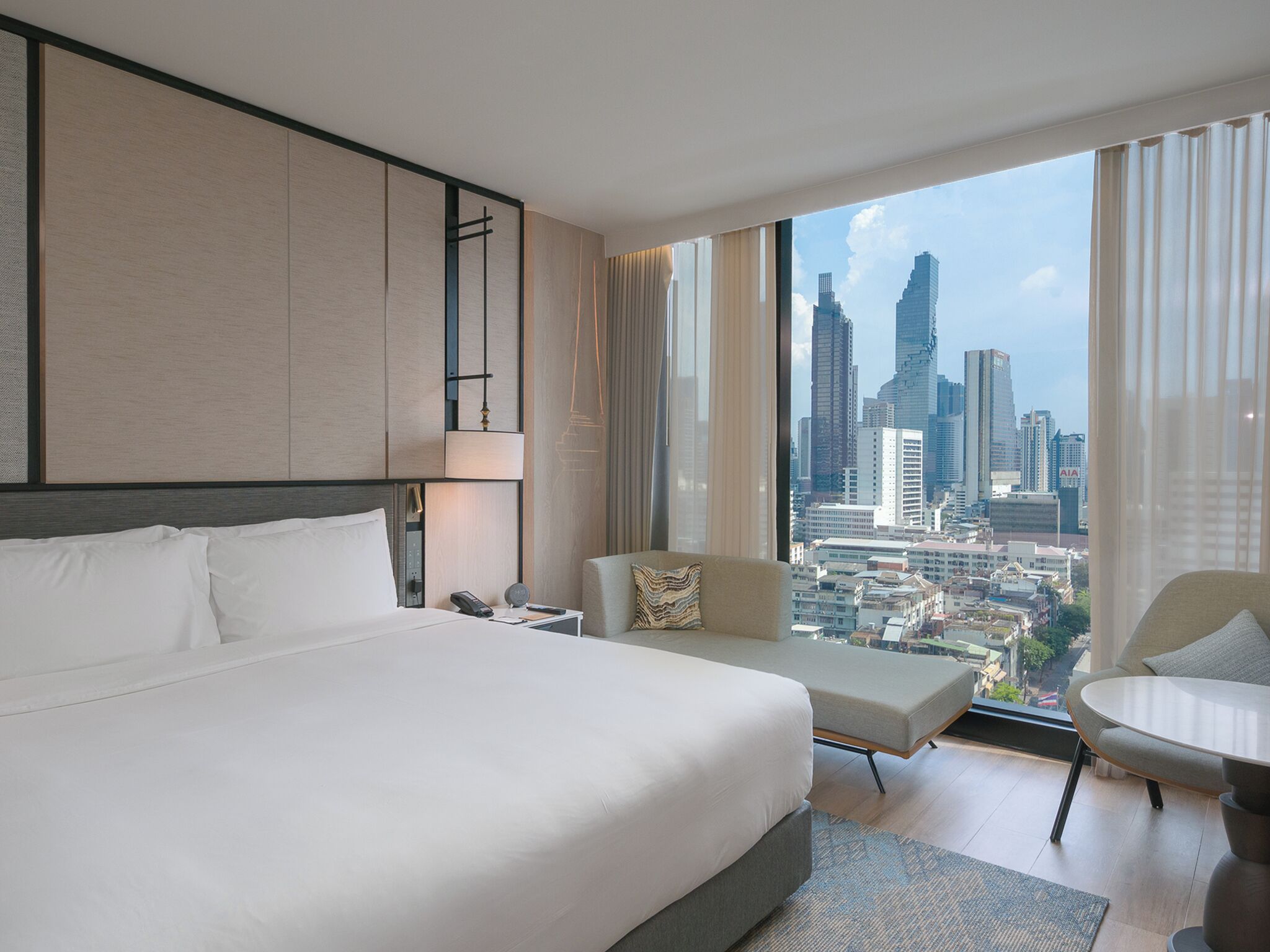 Habitación de hotel con una cama grande, una silla, una mesita y vistas a la silueta de la ciudad.