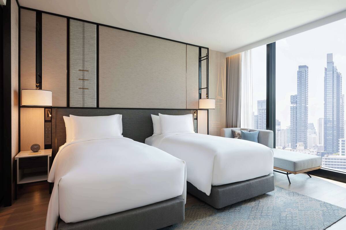 A hotel room with two beds, a nightstand, a chair, and a large window with a city view.