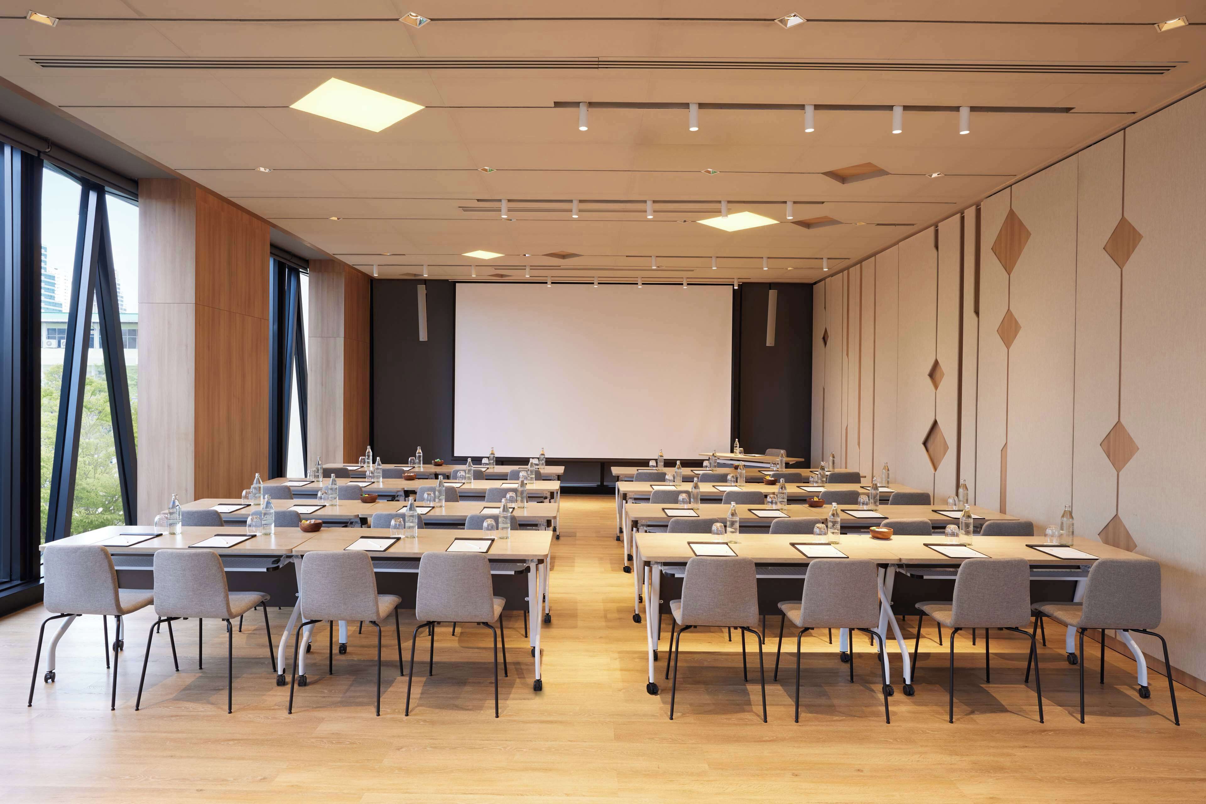 Un salón de conferencias con varias mesas y sillas, una pantalla de proyector y ventanas grandes.