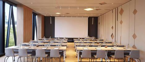 Un salón de conferencias con varias mesas y sillas, una pantalla de proyector y ventanas grandes.