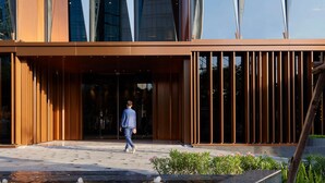 Un edificio moderno con fachada de vidrio, entrada de madera y zona ajardinada con plantas.