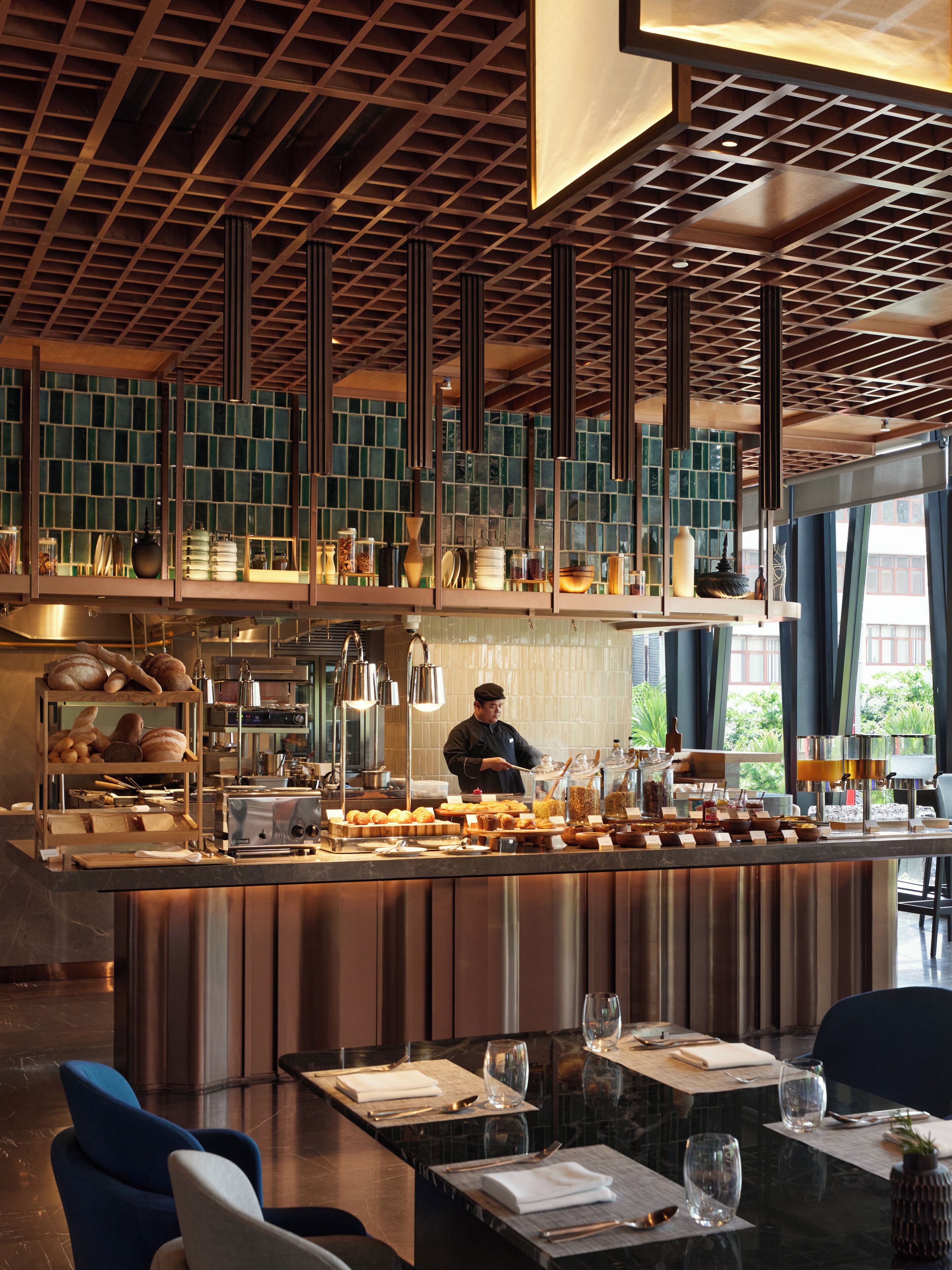 Una cocina moderna con un chef preparando comida, una zona de bar con diferentes opciones gastronómicas y un comedor con mesas listas para los comensales.