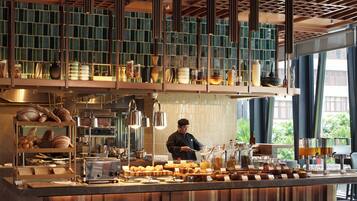 Una cocina moderna con un chef preparando comida, una zona de bar con diferentes opciones gastronómicas y un comedor con mesas listas para los comensales.