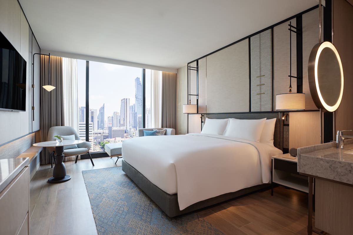 A modern hotel room with a large bed, a desk, a chair, and a view of the city skyline.