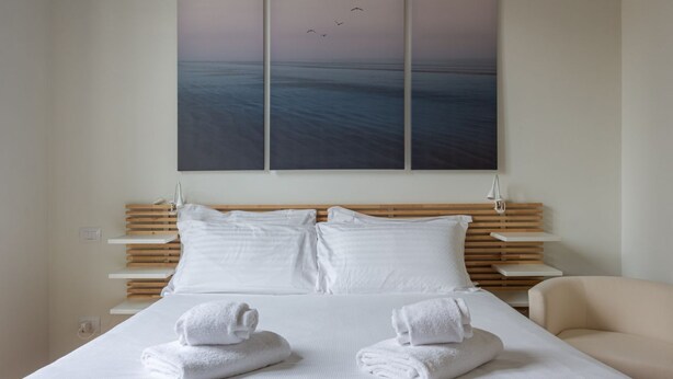 A hotel room with a bed, white linens, two pillows, a wooden headboard, a wall-mounted artwork, and a beige armchair.