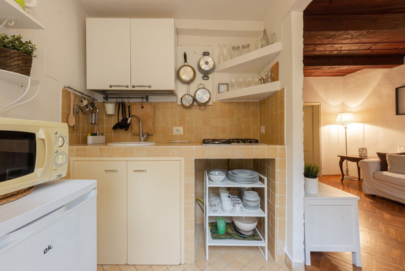 Cocina compacta con armarios blancos, microondas y un refrigerador pequeño. Se observan estantes con vajilla y un colgador para ollas.