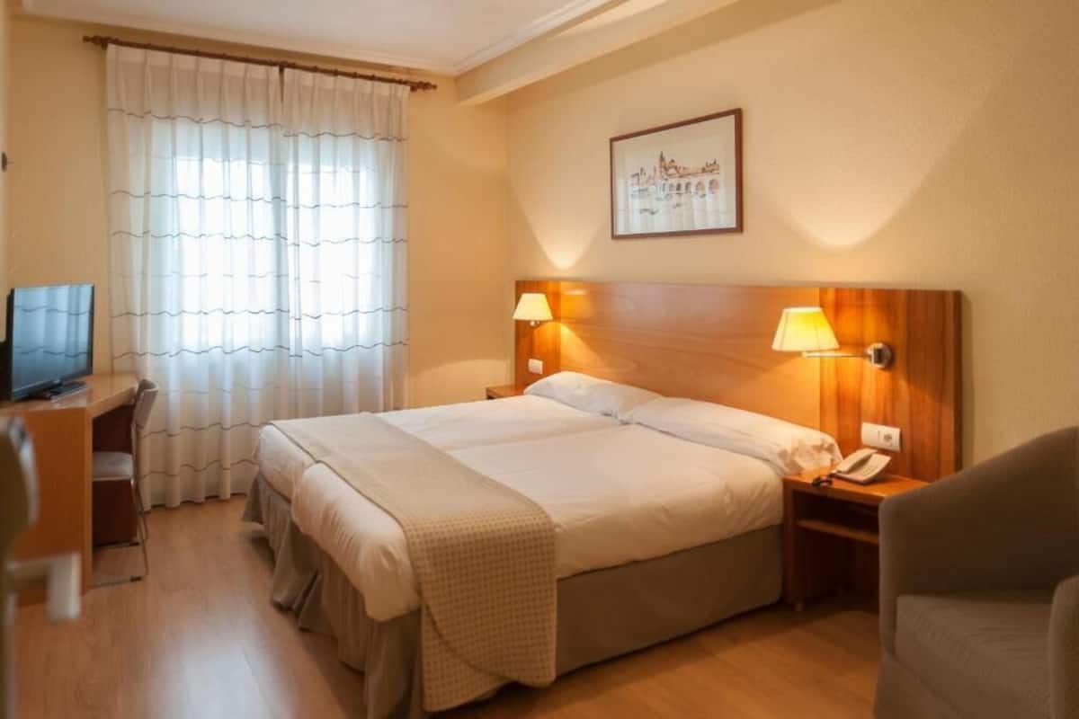 A hotel room with a double bed, two bedside lamps, a desk with a chair, a television, and a framed picture on the wall.