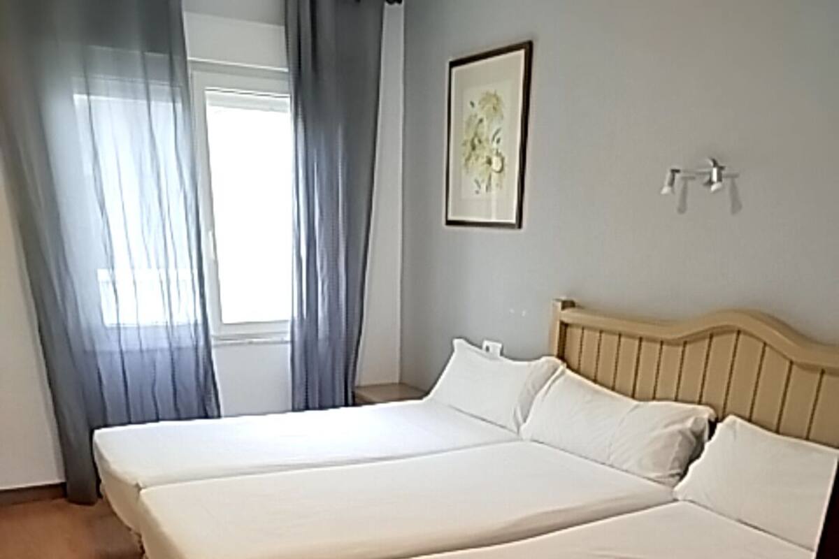 A room with two single beds, a wooden floor, a window with curtains, and a framed picture on the wall.