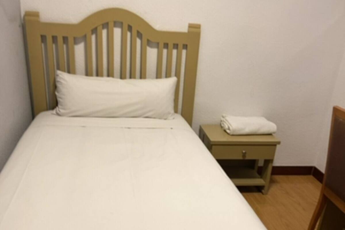 A single bed with a wooden headboard, a small bedside table with a folded white towel, and a chair in a room with wooden flooring and plain walls.