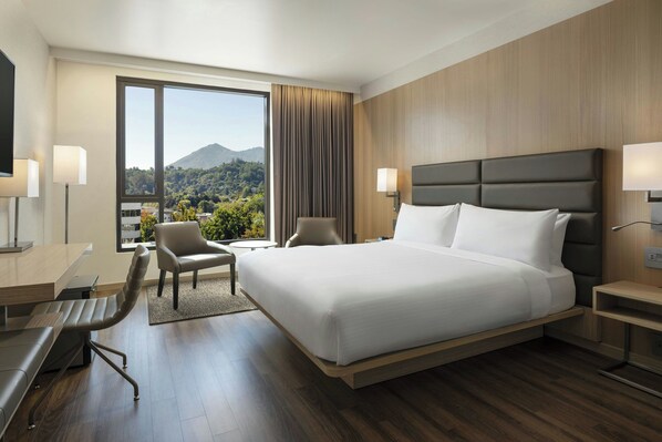 A modern hotel room with a large bed, a desk, and a chair. There is a view of a mountain and trees through the window.