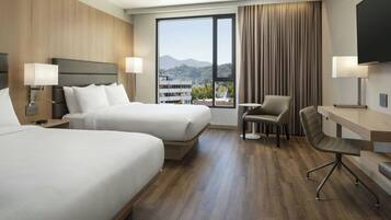 Habitación de hotel con dos camas, un escritorio, una silla y un ventanal con vistas a edificios y montañas.