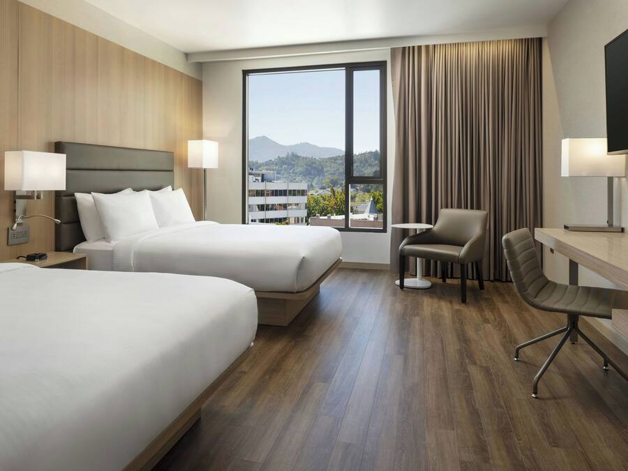 Habitación de hotel con dos camas, un escritorio, una silla y un ventanal con vistas a edificios y montañas.