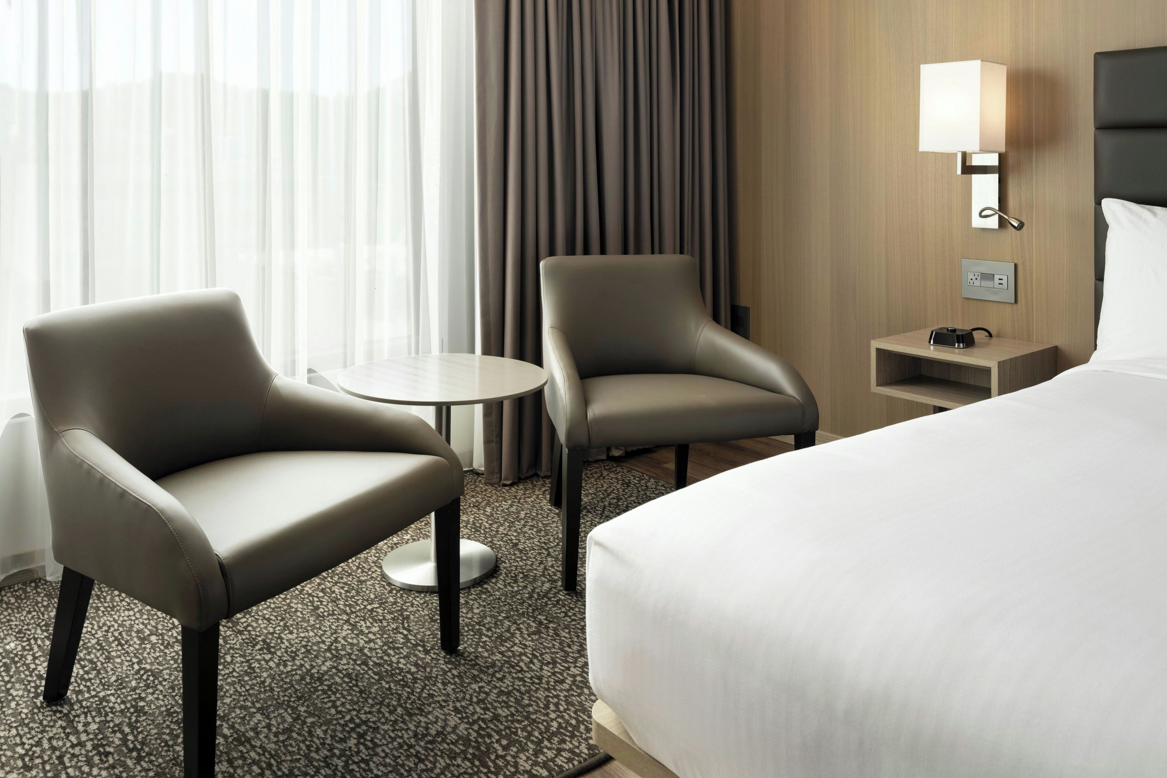 A hotel room with a bed, two armchairs, a small table, a lamp, and a nightstand.