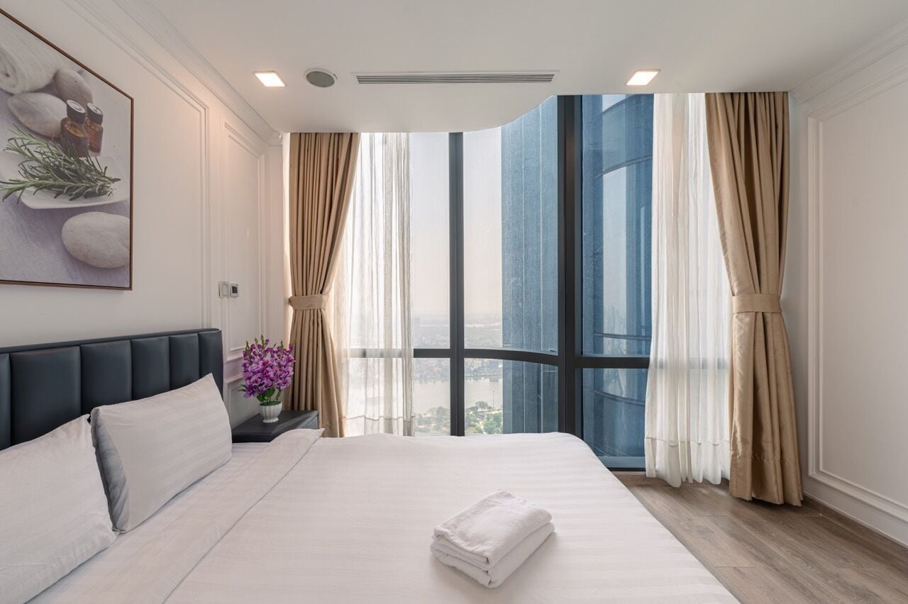 Une chambre d’hôtel avec un grand lit, une table de chevet ornée d’un vase de fleurs, un tableau au mur et une vue sur la ville à travers de grandes fenêtres.
