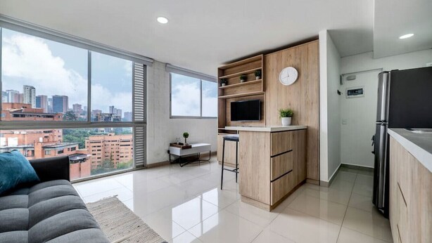 A modern apartment with a kitchen, a living area with a sofa and a TV, and large windows offering a city view.