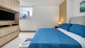 A modern bedroom with a large bed, a TV on a wooden cabinet, a window with a city view, and a nightstand with a lamp.
