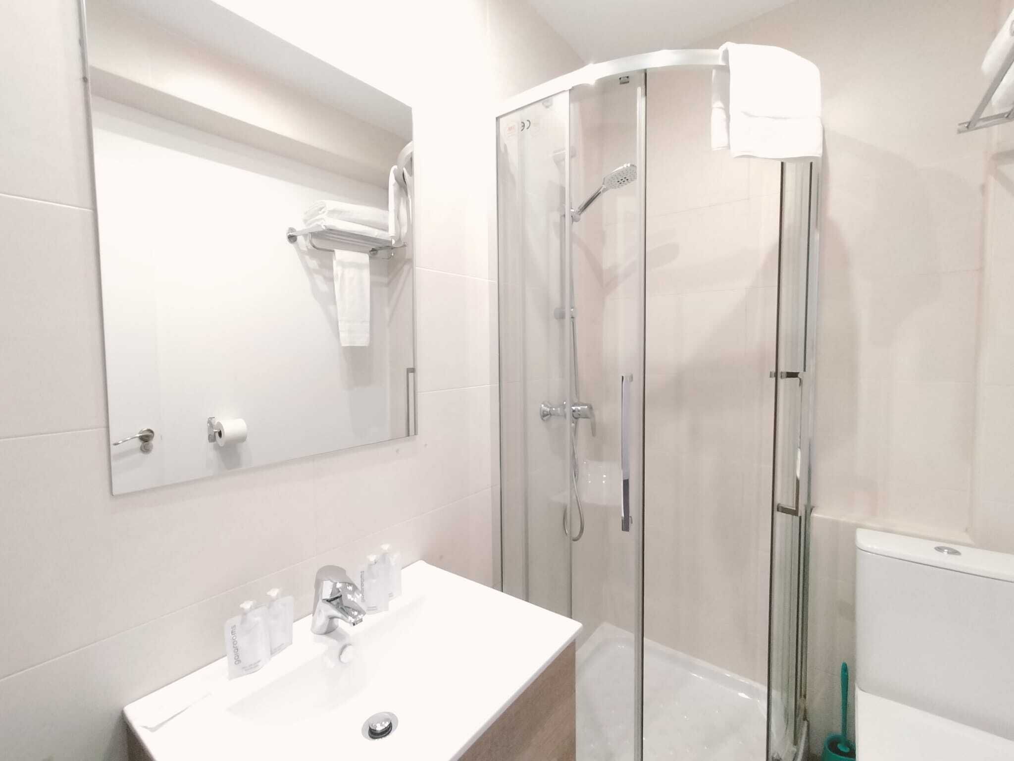 Un baño moderno con ducha de vidrio, un lavamanos blanco con espejo arriba y un inodoro a la vista en la esquina.