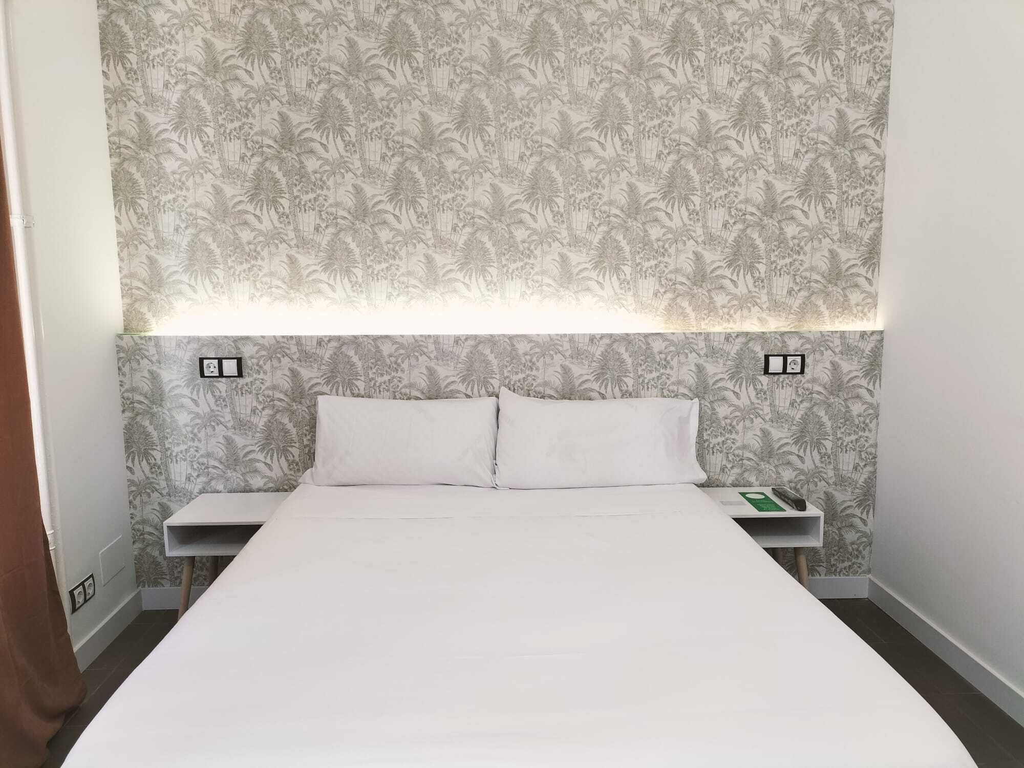 Habitación de hotel con una cama grande, dos mesitas de noche y un cabecero con estampado de palmeras. Se observan dos interruptores de luz en la pared y un objeto verde sobre la mesa de la derecha.