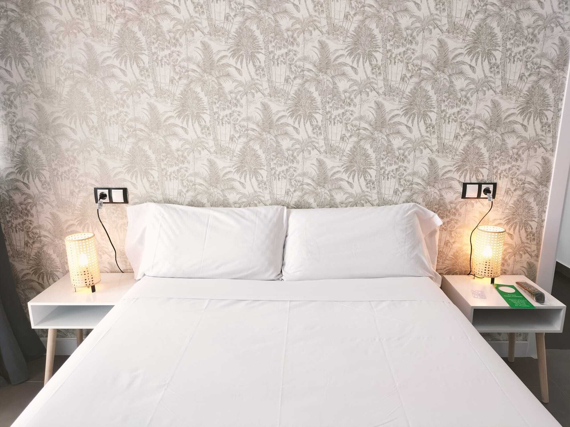 Una cama con ropa blanca, dos mesitas de noche con lámparas y una pared con estampado de palmera tropical.