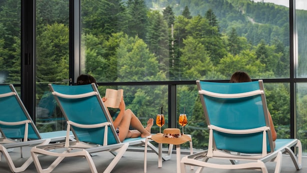 A person relaxing on a balcony chair with a view of a forested mountain.