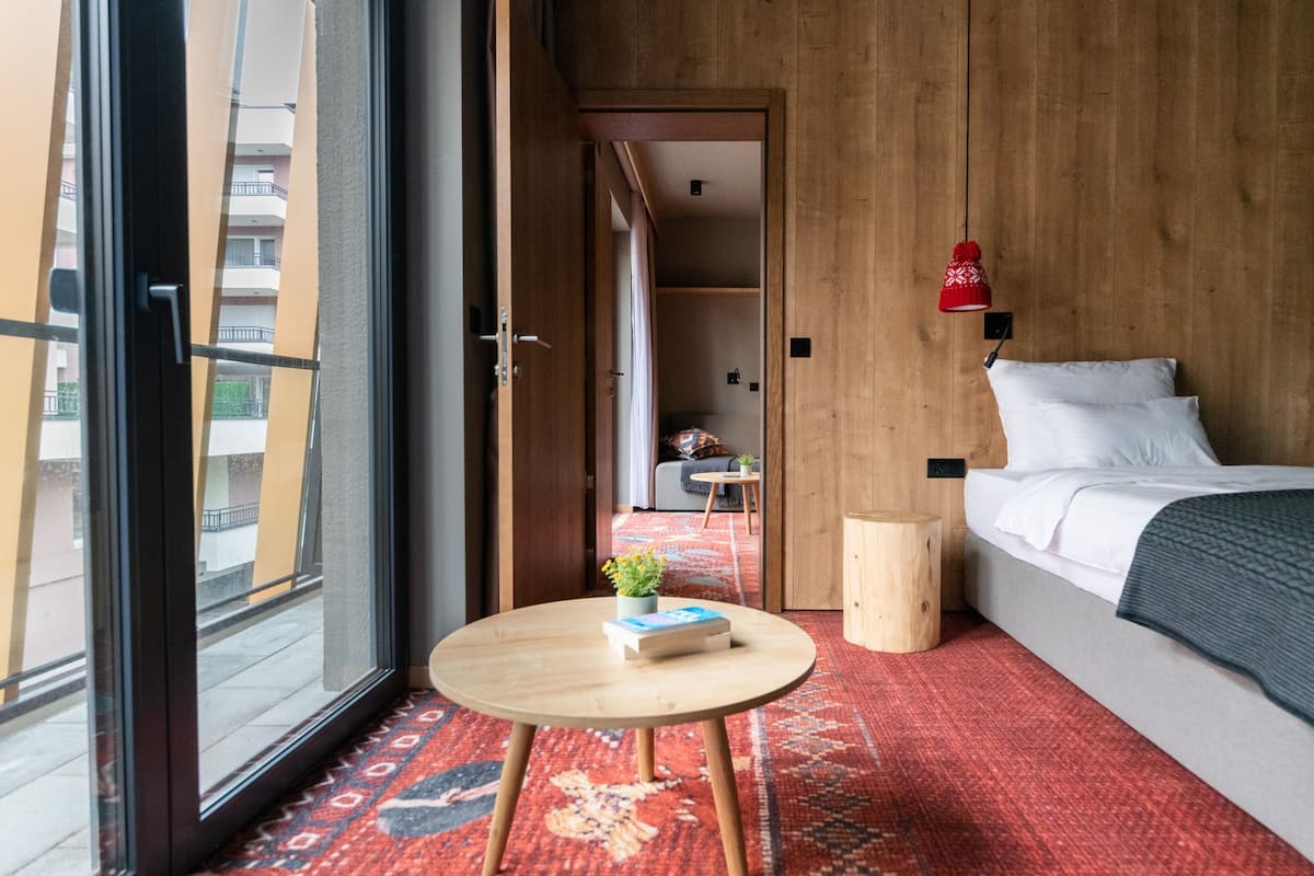 Een hotelkamer met een groot raam, een bed met witte lakens, een rode lamp en een klein rond tafeltje met een boek en een plant.