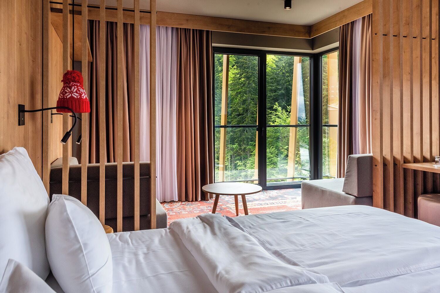 Habitació d'hotel moderna amb un llit gran, una taula rodona petita i vistes a vegetació a través de grans finestres.