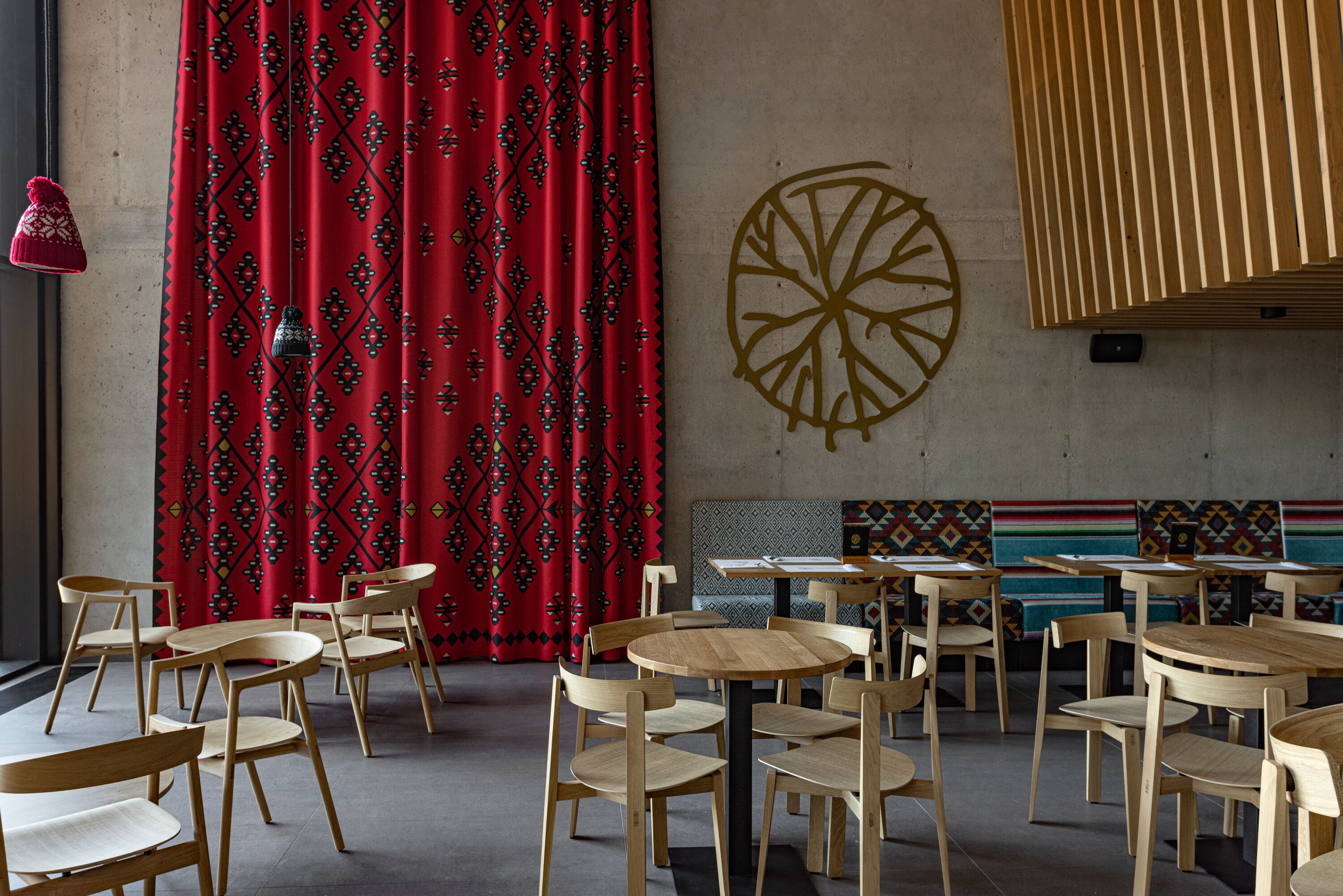 Een restaurantinterieur met ronde houten tafels en stoelen, rode gordijnen met een patroon en een grote wanddecoratie.