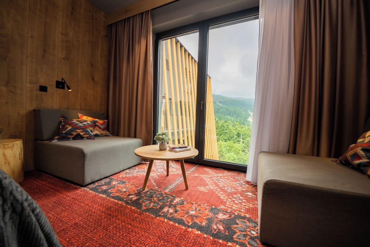 Een moderne kamer met een bank, een klein rond tafeltje en een groot raam met uitzicht op een bebost landschap.