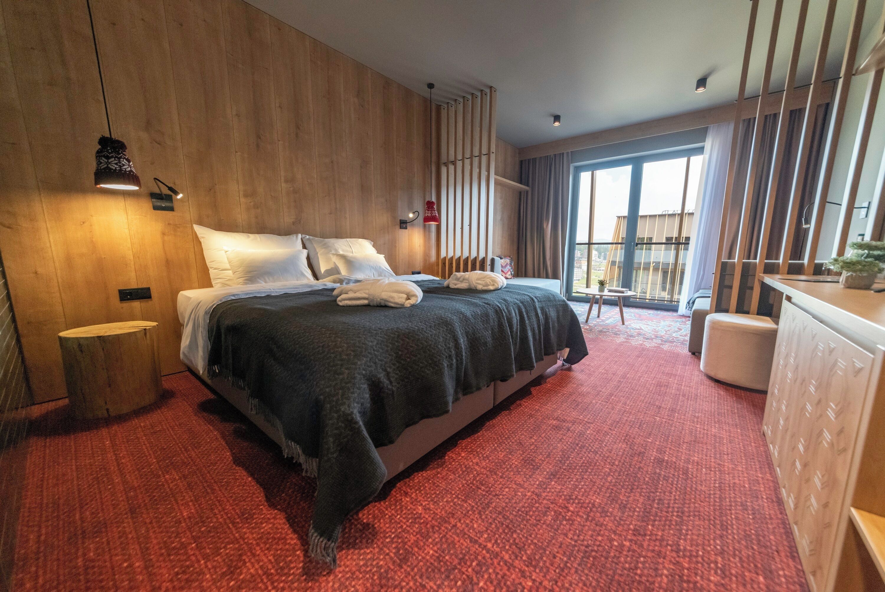 Une chambre d’hôtel moderne avec un grand lit, des murs recouverts de panneaux en bois, une moquette rouge et une vue sur le balcon extérieur.