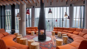 A modern lounge area with orange seating, a central wood-burning stove, and large windows offering a view of the forest.