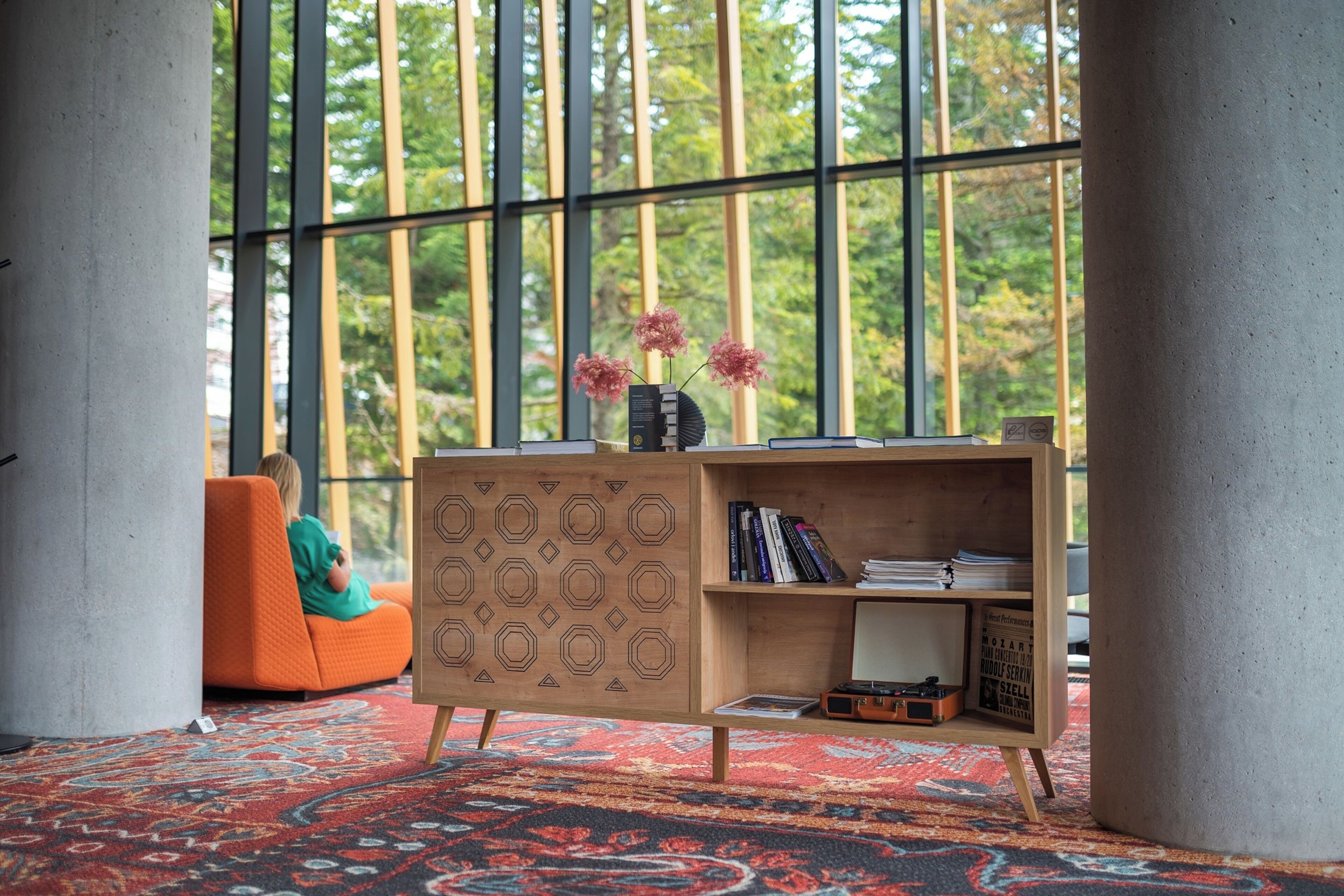A modern room with a large window, a wooden cabinet with books, a patterned carpet, and an orange chair.